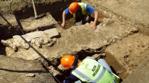 Albion Archaeology Two people in a pipe trench, they are both bending over, so you cannot see their faces. They are wearing orange hard hats, high-vis jackets. There are tools around them and soil. 