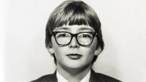 RCS Black-and-white portrait of a young Tennant wearing a formal suit and tie, with straight hair parted near the center, against a plain light background.