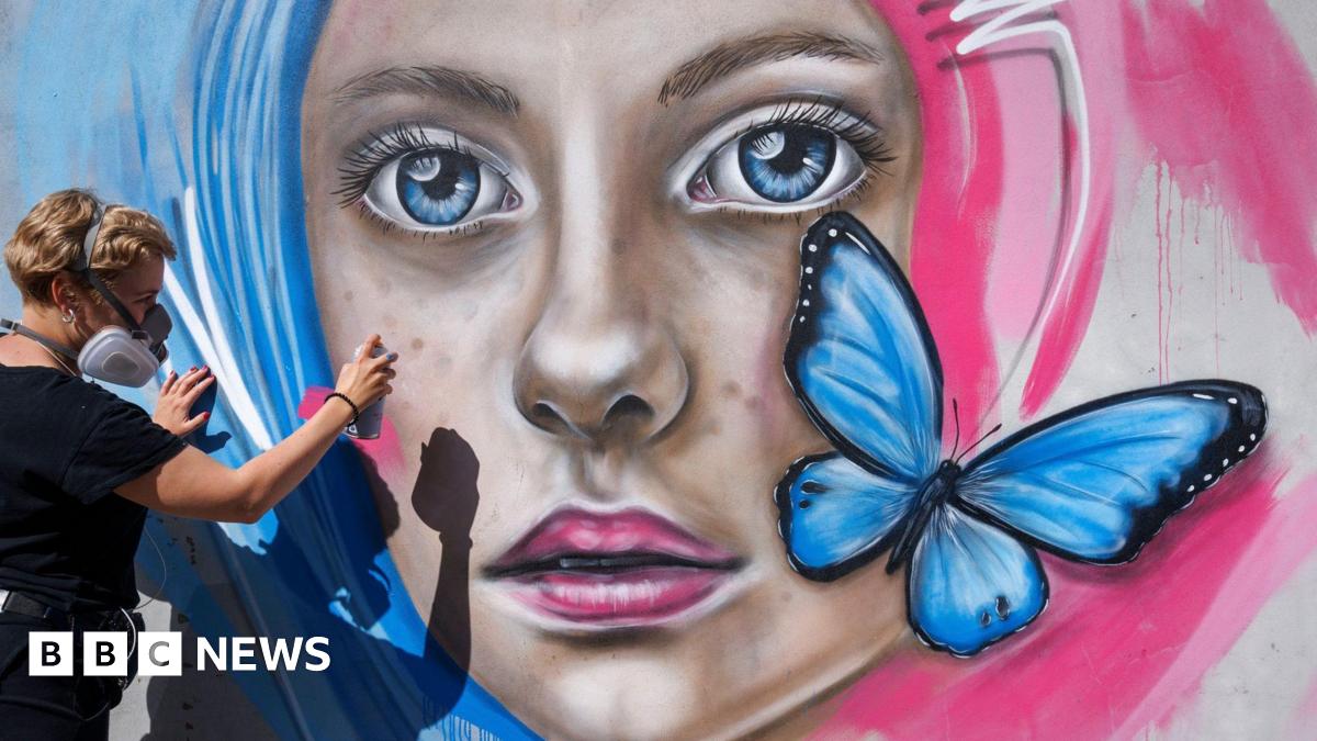 A woman paints a mural of a woman with pink and blue hair. The mural has a blue butterfly on it.