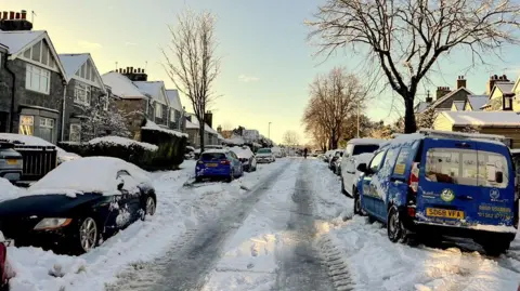 PA Media Snow-covered residential street with parked cars lining both sides, icy road surface, and houses and trees under a clear winter sky