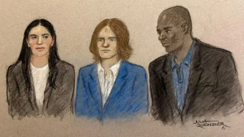 Julia Quenzler A court sketch of Mckenzie Morgan. He is wearing a blue suit and with hair that is longer than in the mugshot, this time reaching his shoulders. He is flanked by two people wearing formal attire.