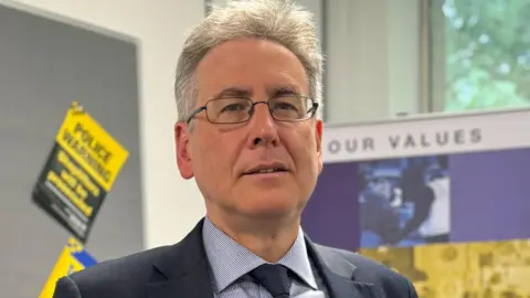 A man with white hair and wire-framed glasses in a room with police leaflets pinned on bulletin boards. He is wearing a navy suit and blue checked shirt.
