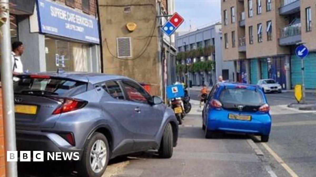 A pavement in a built up area. A grey car is parked fully on the pavement, as is a motorbike with a blue box on the back. A smaller blue car is partly parked on the road and partly on the pavement.