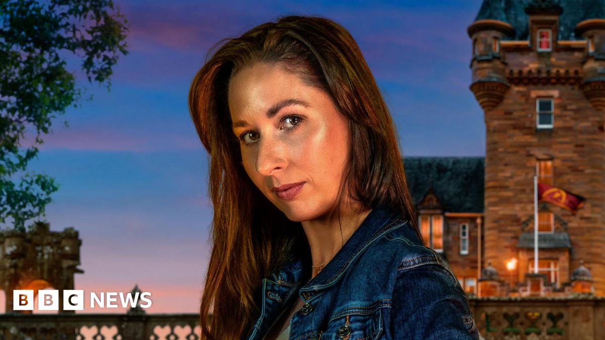 A close-up image of Rachel Duffy from County Down. She is looking at the camera with a neutral expression. She has long, dark hair and is wearing a blue denim jacket. Behind her is the traitors Castle, with the sky getting darker as the sun sets.