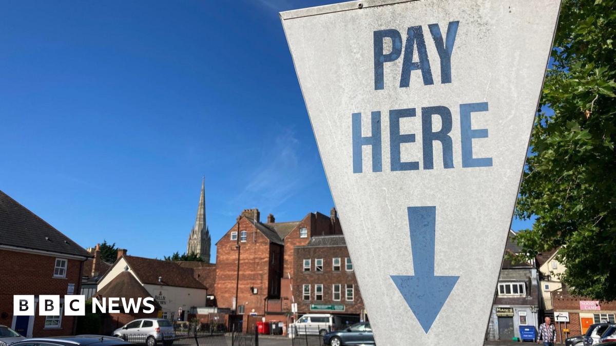 A white sign at a car park in Salisbury, Wiltshire, which reads "pay here" in blue writing, with a blue arrow pointing down. The sign is shaped like an upside down triangle. Cars can be seen parked in the city car park behind, which is surrounded by older buildings and a large spire can be seen in the background. It is a sunny day.