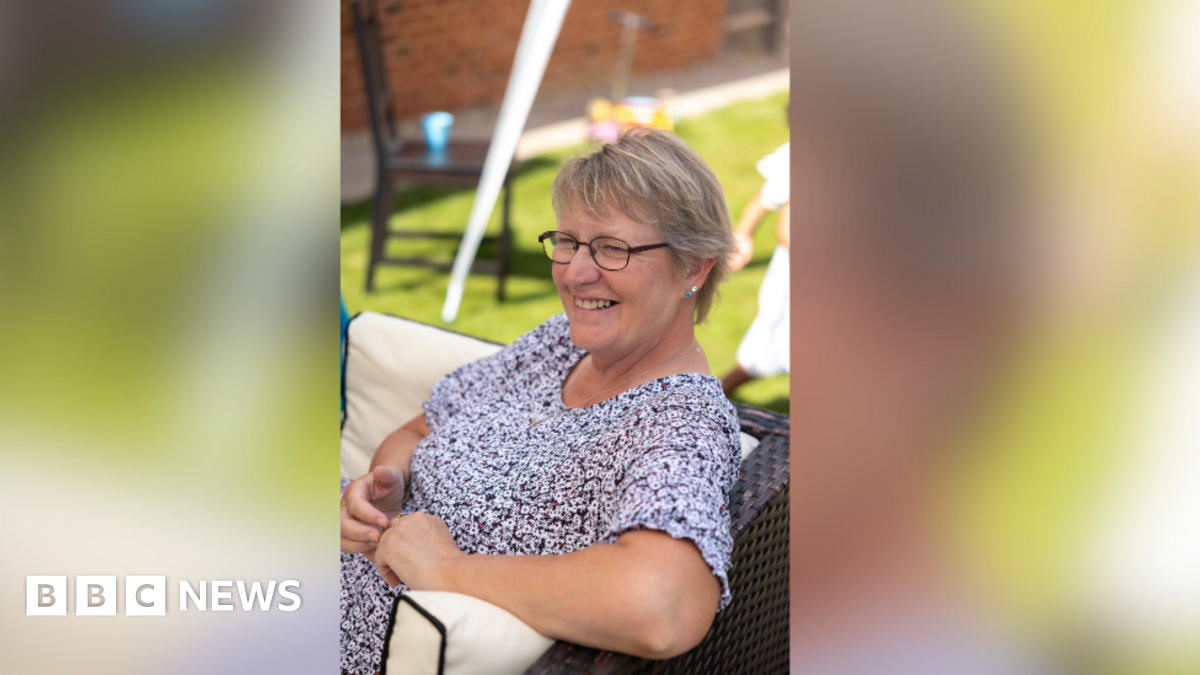 Suzanne Wilmot sits in a garden chair in a sunny garden, she wears a black and white floral top and has short blond hair and glasses. She is smiling at someone out of shot.