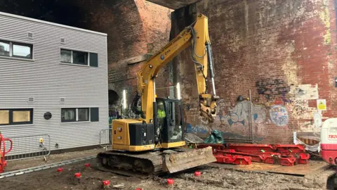Photograph of the Embassy homeless village, which is under construction on the edge of Manchester city centre. The image includes a digger on a construction site underneath railway arches.