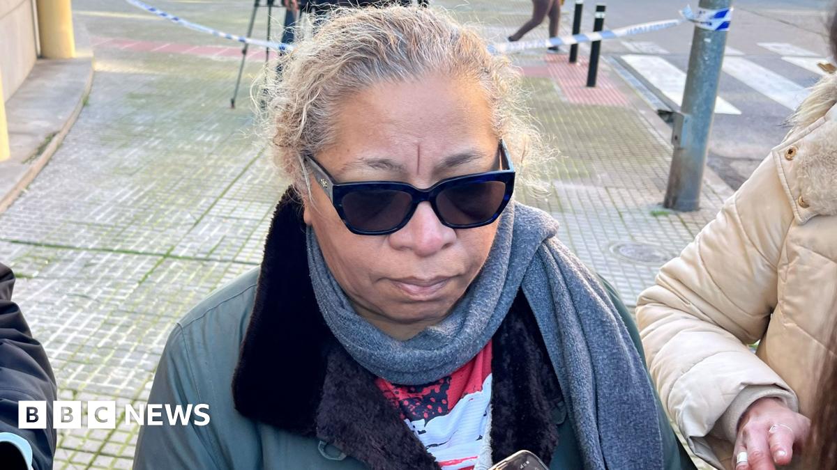 A woman in a grey scarf and coat talks to reporters