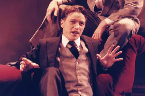 RCS McAvoy dressed in a formal three-piece suit with a tie, seated on a red upholstered chair with hands raised in an expressive gesture. Another person’s legs, also in formal attire, are visible resting on the arm of the chair. The background has a warm reddish tone, suggesting a theatrical or staged setting