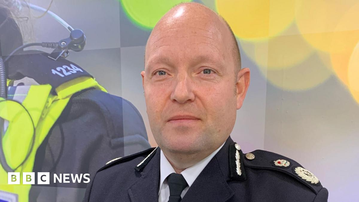 Millie Bobby Brown and David Harbour in character as Eleven and Jim Hopper. Eleven wears red shorts over grey leggings and a grey jumper. Jim Hopper wears a black leather jacket, a black beanie hat and dark trousers.They are speaking to director Frank Darabont on the set of Stranger Things. There is a vintage advertisement posted in the background with debrief on the ground.