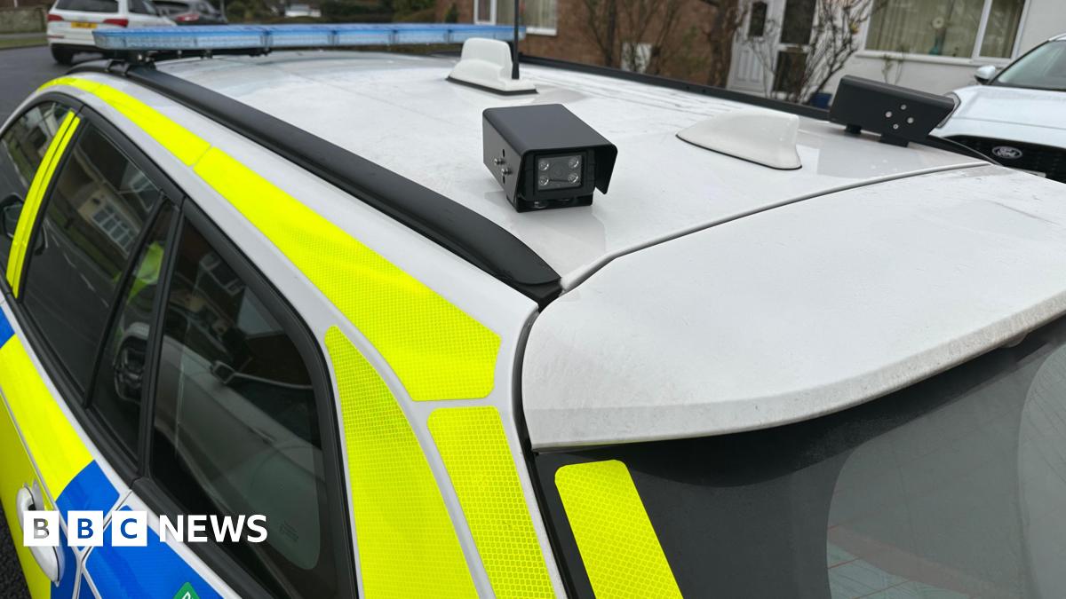 Millie Bobby Brown and David Harbour in character as Eleven and Jim Hopper. Eleven wears red shorts over grey leggings and a grey jumper. Jim Hopper wears a black leather jacket, a black beanie hat and dark trousers.They are speaking to director Frank Darabont on the set of Stranger Things. There is a vintage advertisement posted in the background with debrief on the ground.