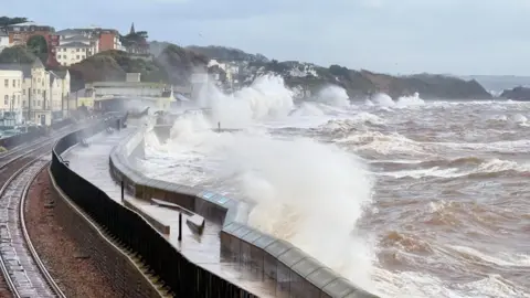 Weather Watchers/AstroPostie The picture shows huge waves crashing over a sea wall during rough weather. Brown, churning water is surging in from the right, and the force of the waves is sending spray high into the air. A railway line runs along the left side of the image, curving around the bay, with wet tracks glistening from the spray. Buildings sit close to the shoreline, and rows of houses climb up the hillside in the distance. The sky is grey and overcast, and the whole scene looks stormy and chaotic, with the sea battering the coast.