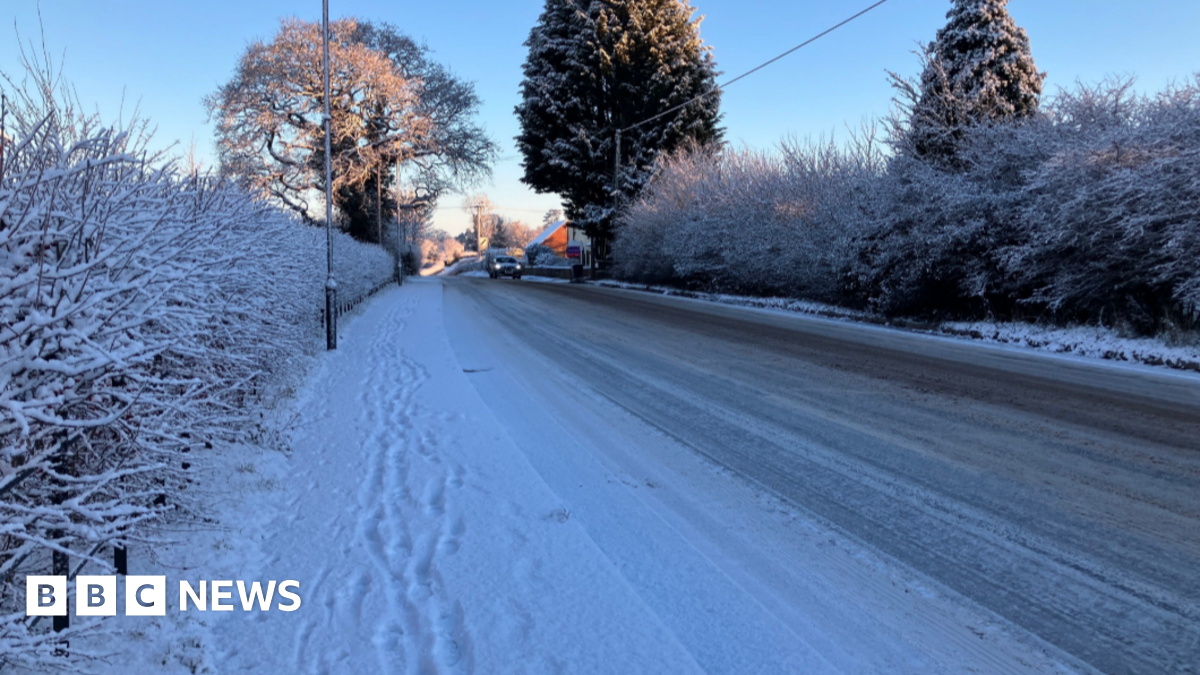 A road and pavement covered in a layer in snow. There is a vehicle approaching with its lights on