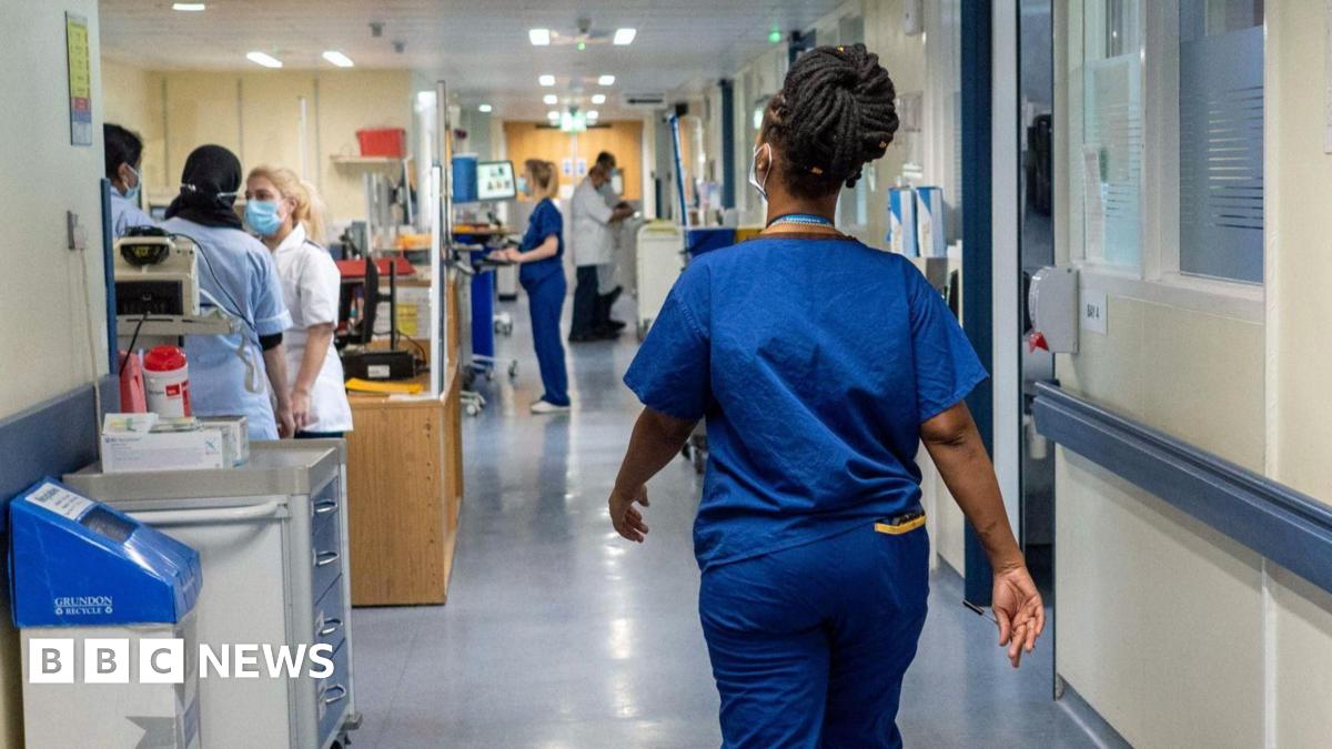 A woman wearing a blue NHS staff uniform walks through the corridor of a hospital away from us.