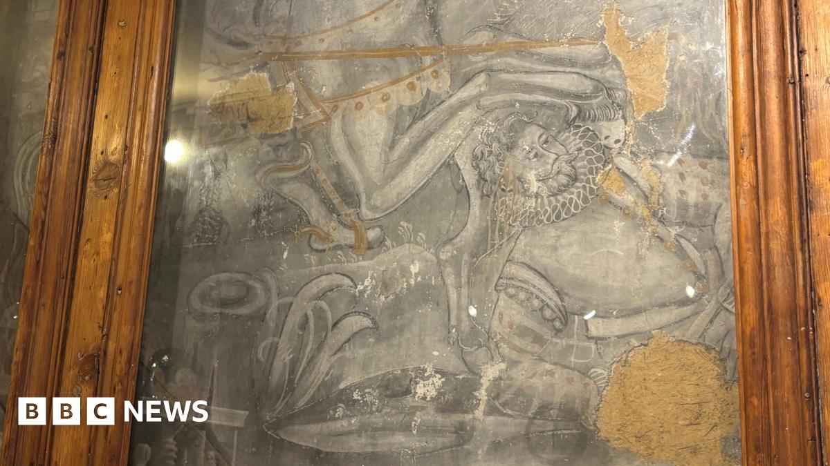 A faded mural on a wall, with wooden frames around it, showing a man on the ground and a horse. The image is hard to make out but is mostly grey and brown, with bits missing.