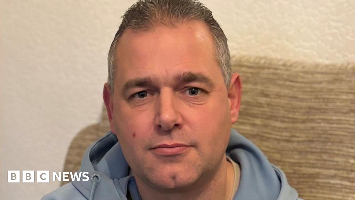 A man in a light blue tracksuit top with short hair sitting on a beige sofa looks at the camera unsmilingly