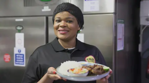 BBC Photograph of Justina John, owner of Onja restaurant. She wears a black polo top which reads "Onja, taste of tanzania" on the yellow circle logo. Her hair is tied up into a black fishnet and she smiles at the camera.
