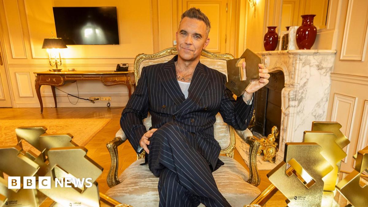 Robbie Williams in a pinstripe suit holding up an Official Charts Company number one trophy and with numerous others on tables either side of him