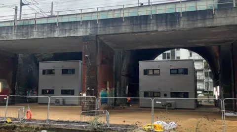 BBC Photograph of the modular homes which are being built underneath a serires of railway arches in Castlefield. The Bridgewater Canal can be seen in the foreground, surrounded by metal fencing.