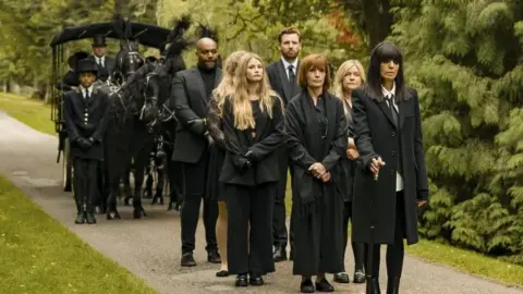 Funeral cortege with Claudia Winkleman leading Traitors contestants including Diane dressed all in black, walking in front of a horse-drawn hearse