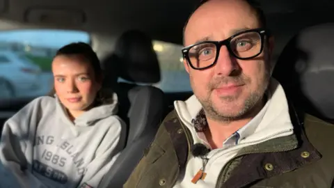 Peter Carmichael is sitting inside a car on the front passenger seat. A woman is in the background in the driver's seat. He has black rimmed glasses and facial hair. He is wearing a green jacket over a cream coloured zip up top.