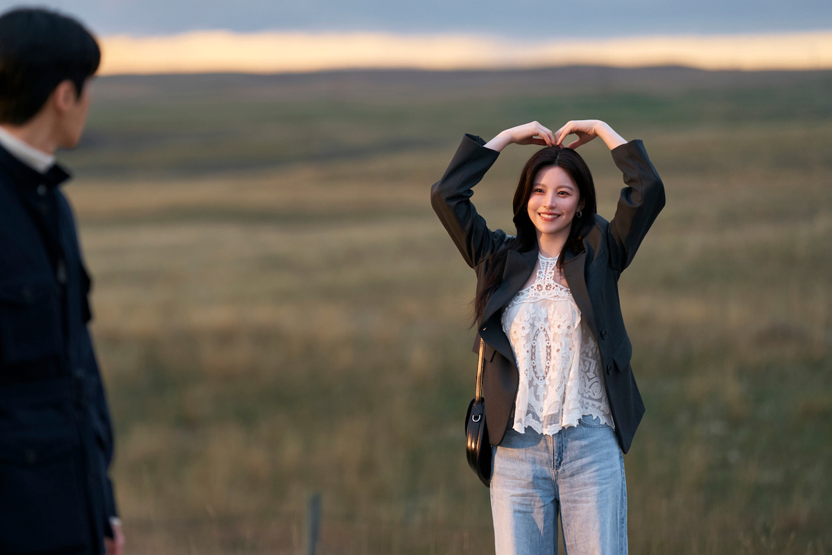 A woman in a casual outfit stands in an open field at sunset, smiling and making a heart shape with her arms above her head, while a man watches her from the side.