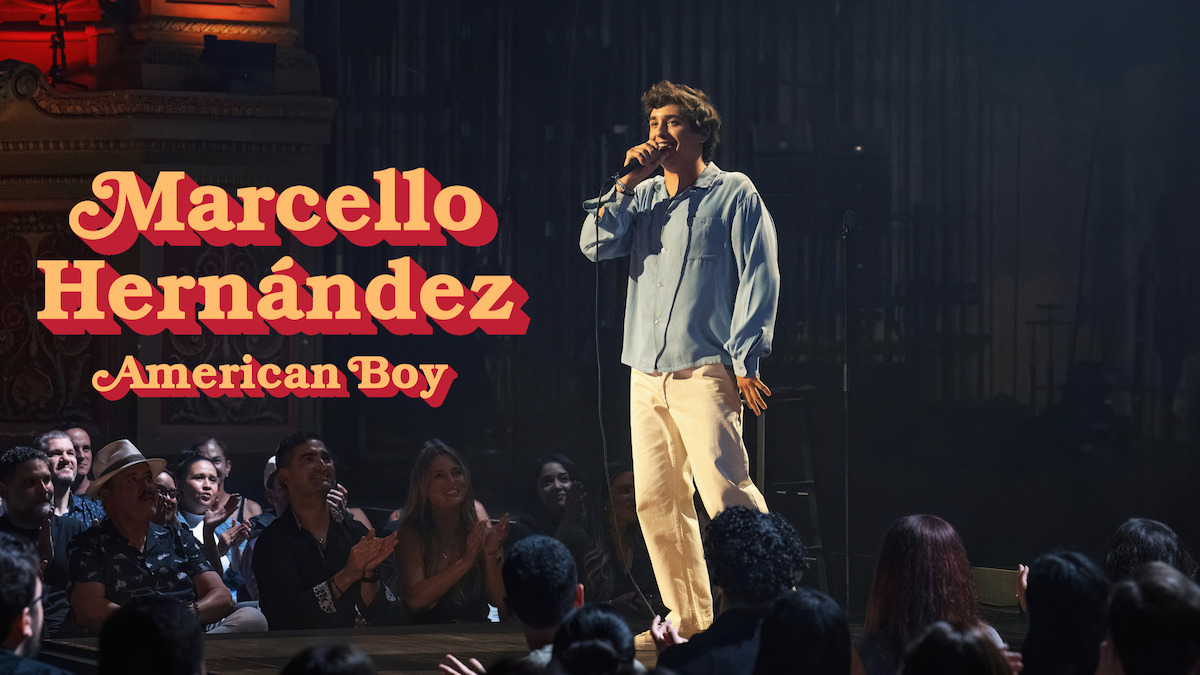 Comedian Marcello Hernández performs on stage in front of an enthusiastic audience in a theater setting, under warm lighting, evoking an energetic and lively mood. Title text "Marcello Hernández: American Boy" appears beside him.