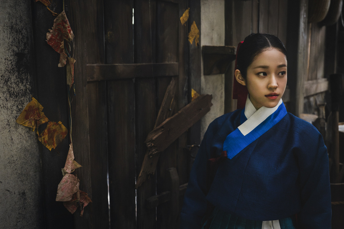 Young woman in traditional Korean hanbok stands near a rustic wooden door and weathered wall, with dried leaves hanging nearby, in a dimly lit, historical setting.