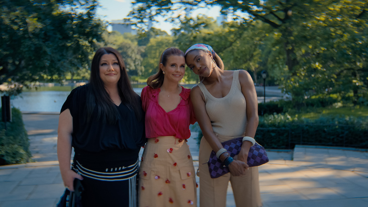 Three women standing close together outdoors on a sunny day in a lush park, smiling and posing for a photo with trees, greenery, and a body of water in the blurred background.