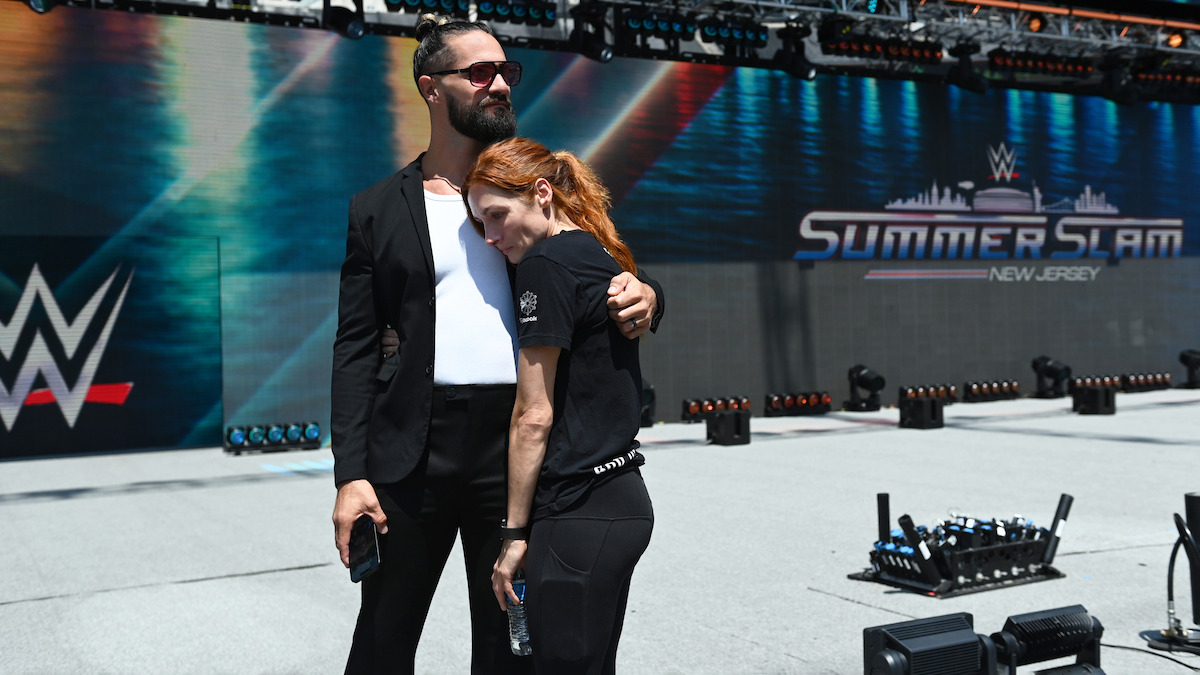 Two people embrace in a staged outdoor arena with SummerSlam and WWE logos in the background, under bright sunlight, conveying a calm and supportive mood before a wrestling event.