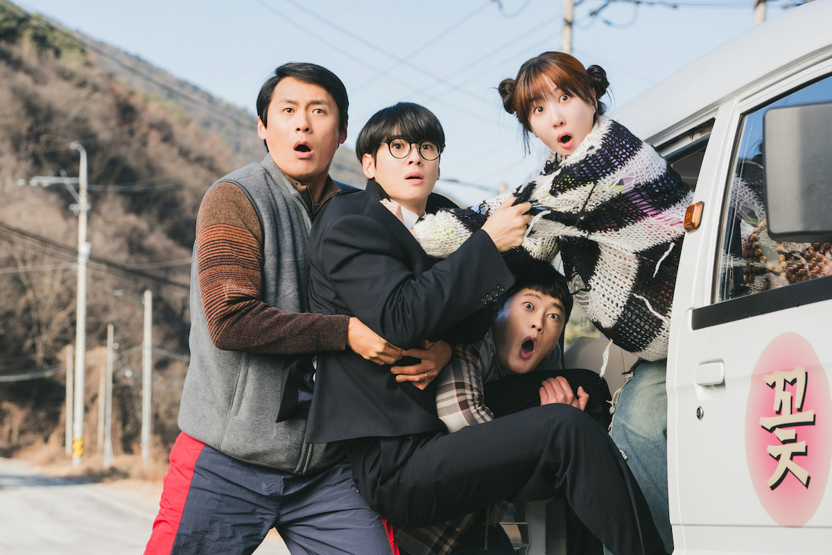 Four surprised people, including a man in a suit and a woman with pigtails, are gathered near a white van on a rural road with hills and power lines in the background, all reacting to something off-camera.