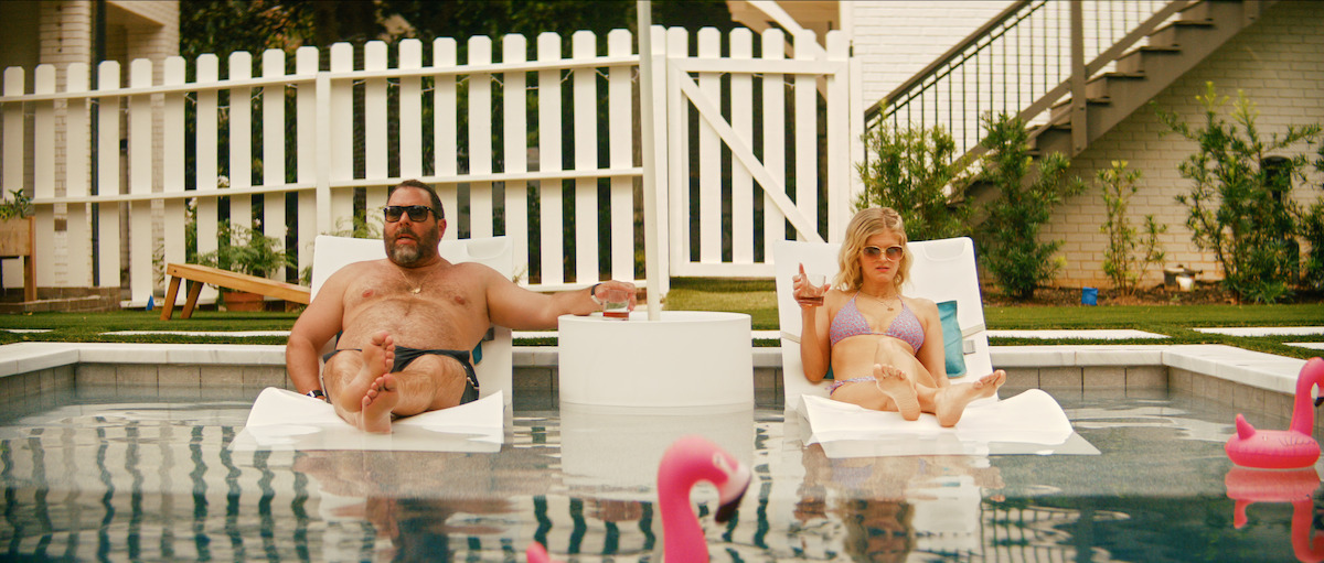 A man and woman relax on pool loungers in a backyard pool, holding drinks. Pink inflatable flamingos float nearby. The pool is surrounded by a white picket fence, green grass, and a two-story house with outdoor stairs.
