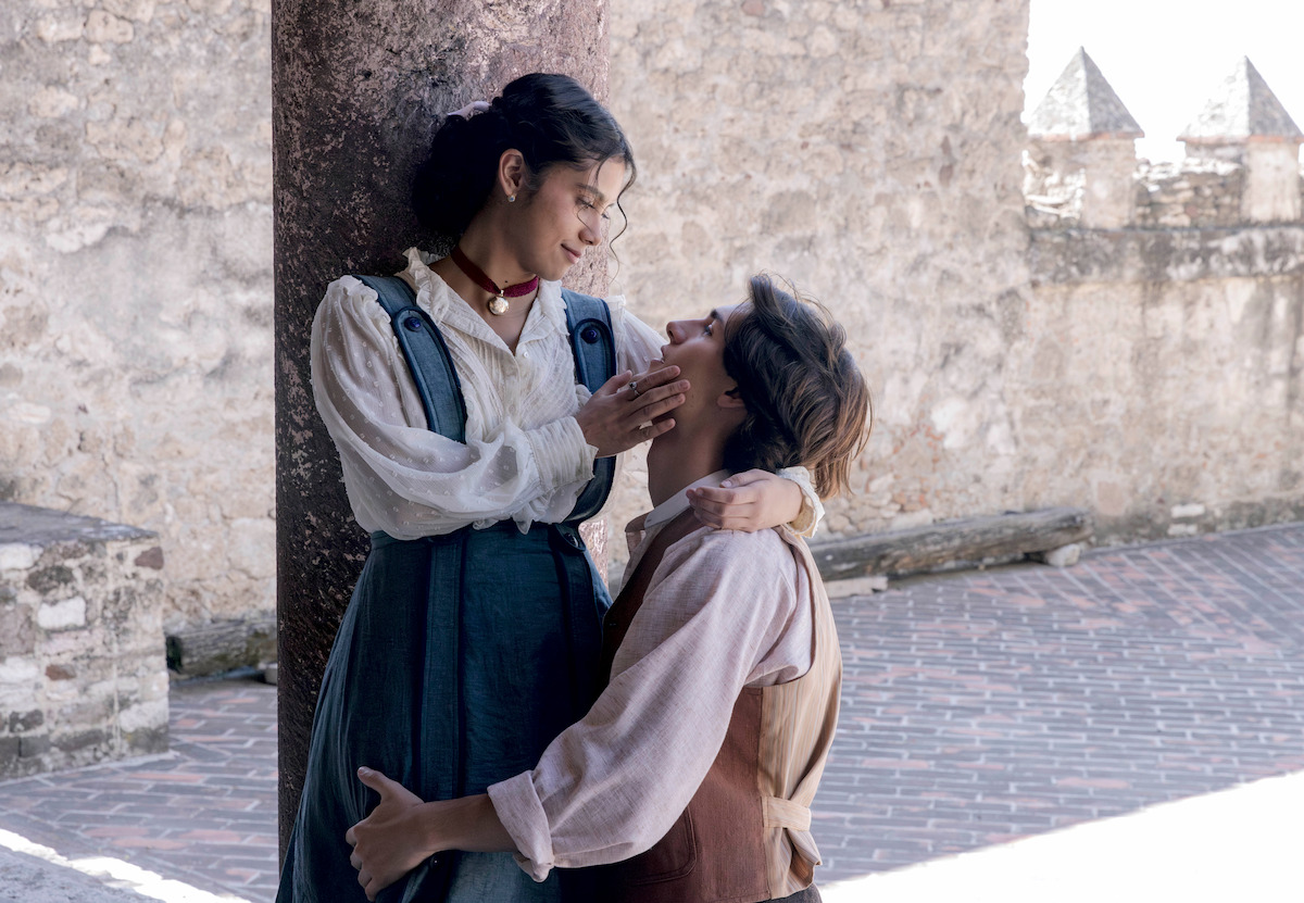 Two people in vintage clothing share an intimate moment outdoors, standing by a stone pillar in a sunlit courtyard with old stone walls and brick pavement, suggesting a historical or romantic scene.