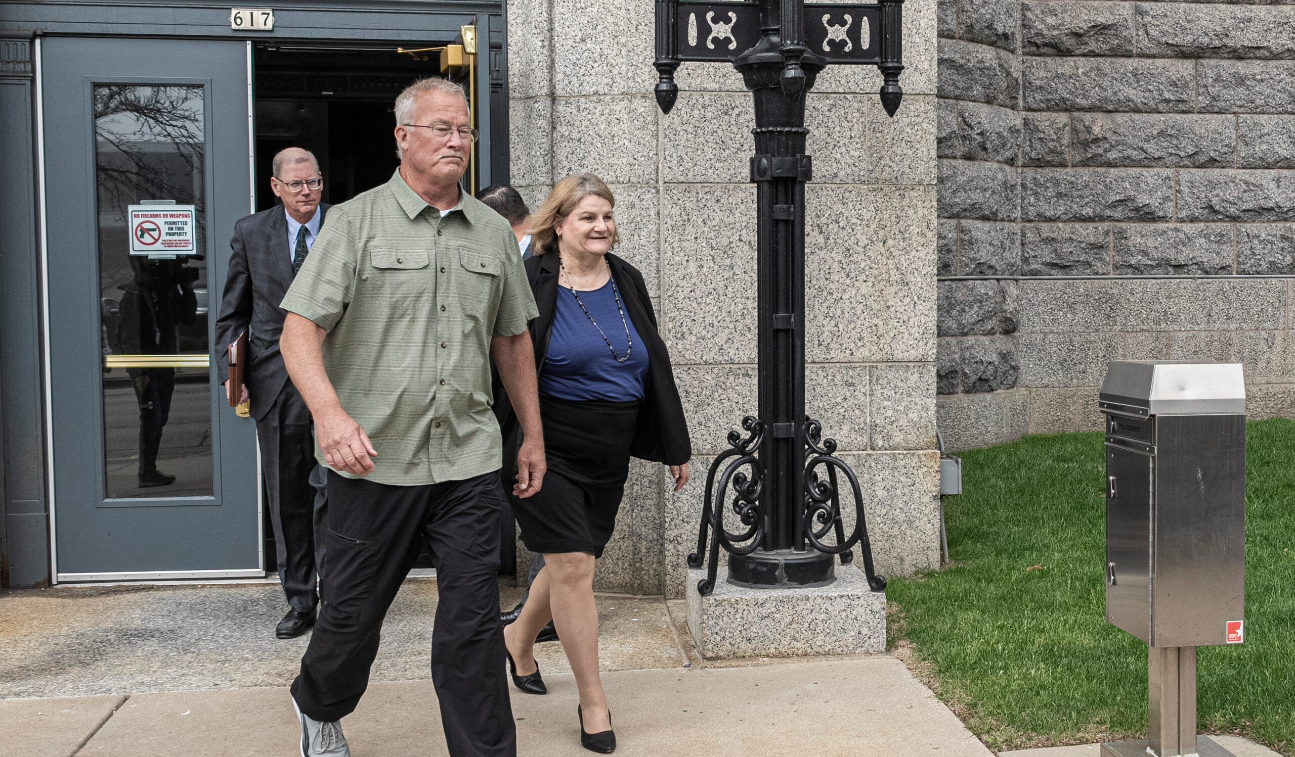 Three adults walk out of a building with stone walls, one man in front in casual clothes, a woman in business attire behind him, and another man in a suit exiting the door.