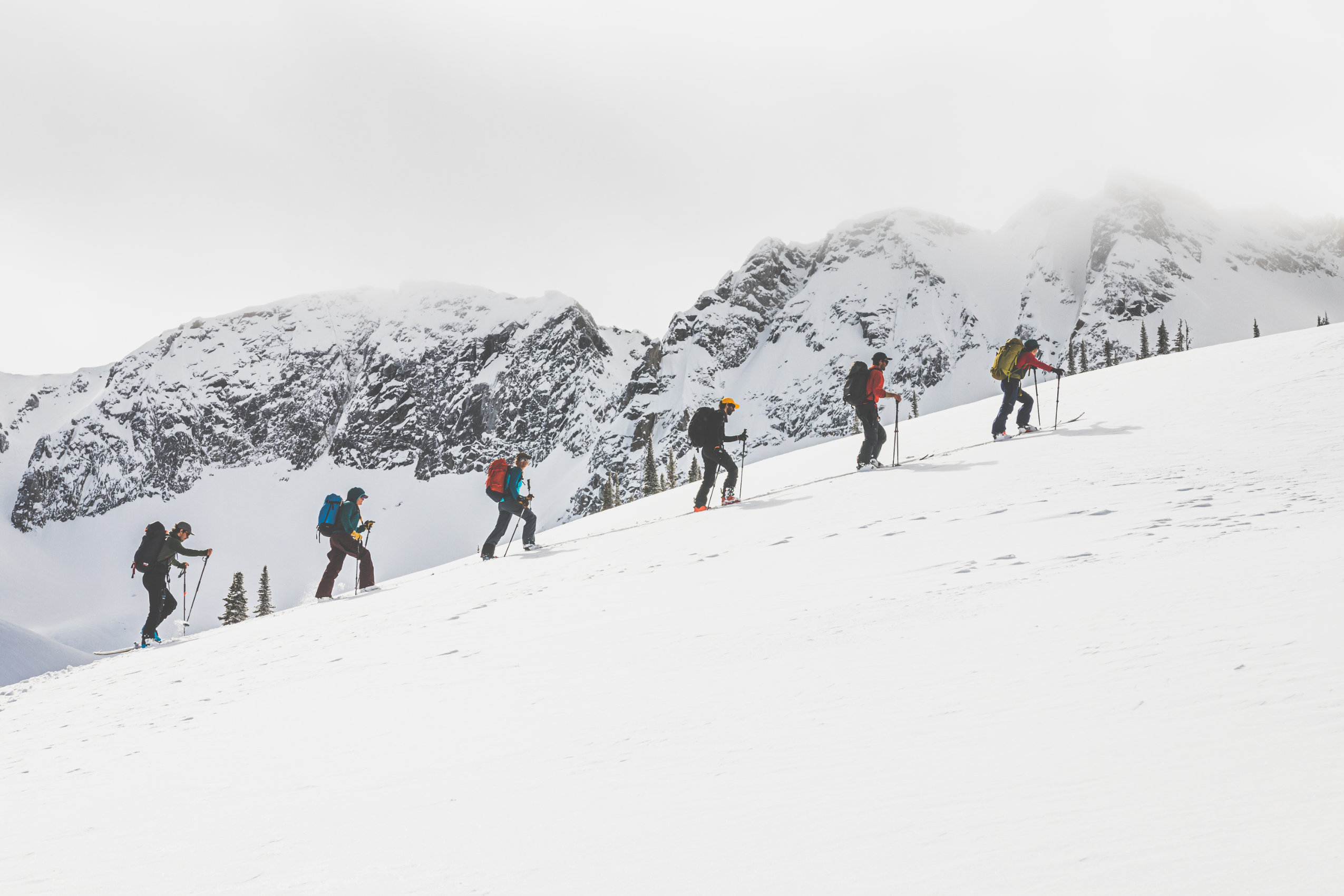 Six skiers in single file climb up a snowy slope, with snow-covered mountains in the background.