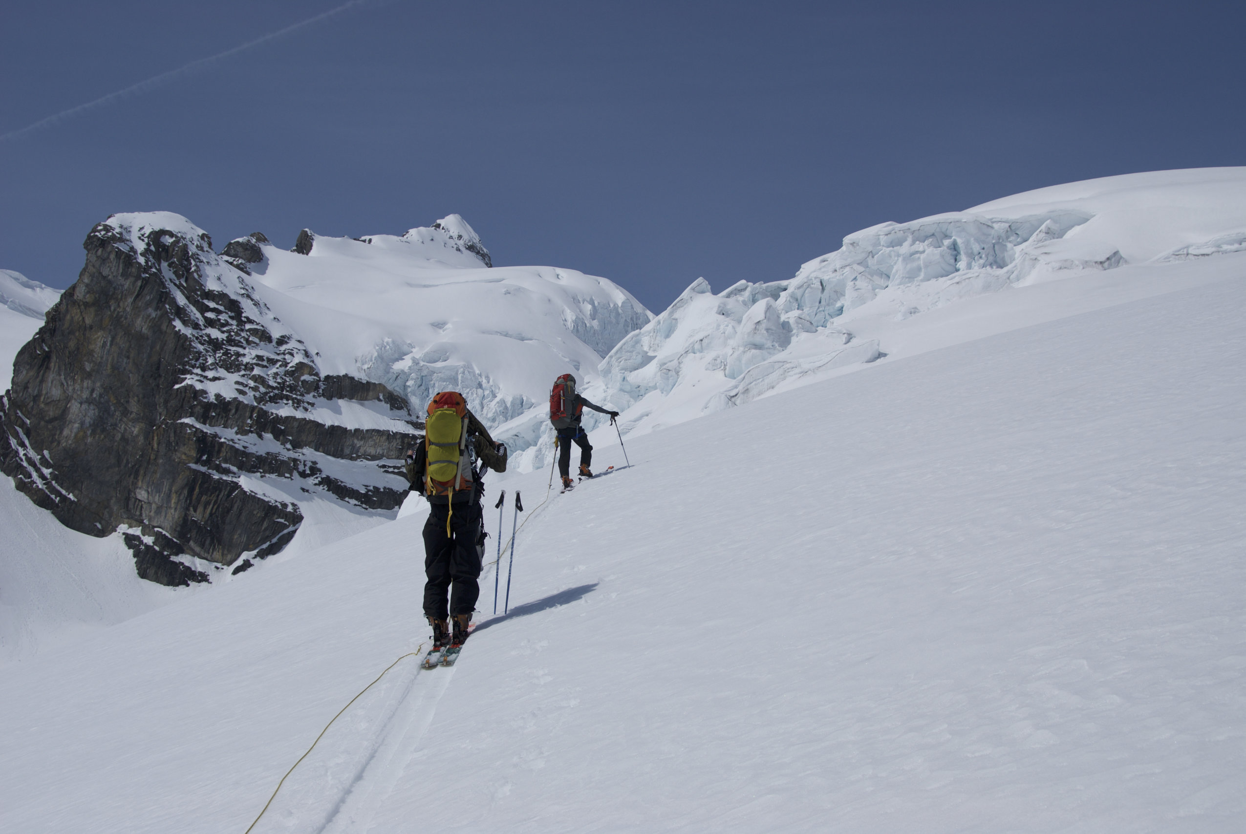 Two alpine explorers climb a snowy slop wearing skies.
