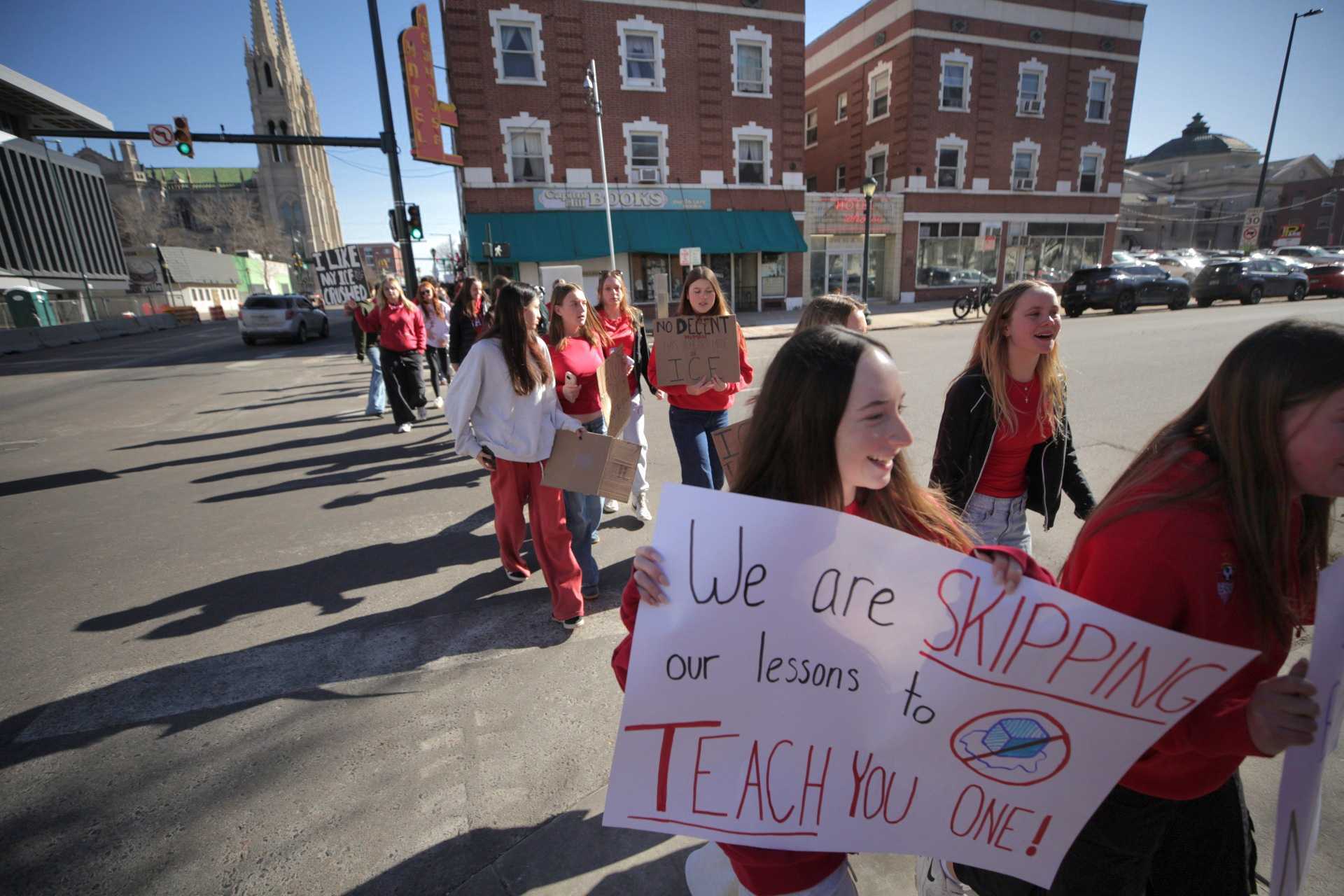 Dozens of high schools students, most wearing red shirts or pants, cross Colfax. A brunette girl in the front holds a sign reading: "We are skipping our lessons to teach you one!"