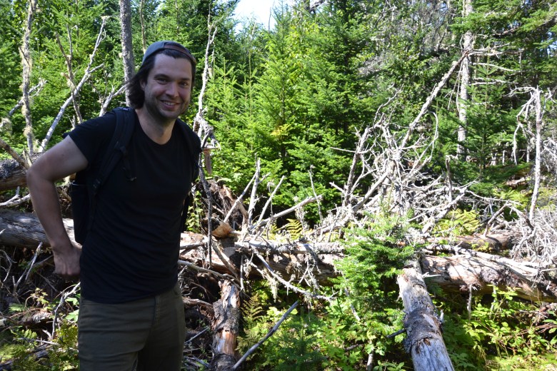 A man in his 30s, wearing a black t-shirt and backwards baseball cap, stands in a forested area, near thick growth and a snarl of fallen trees.