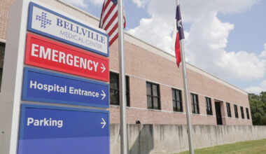 A photo of an emergency room sign for Bellville Medical Center. The American flag and the Texas flag are seen on poles next to it.