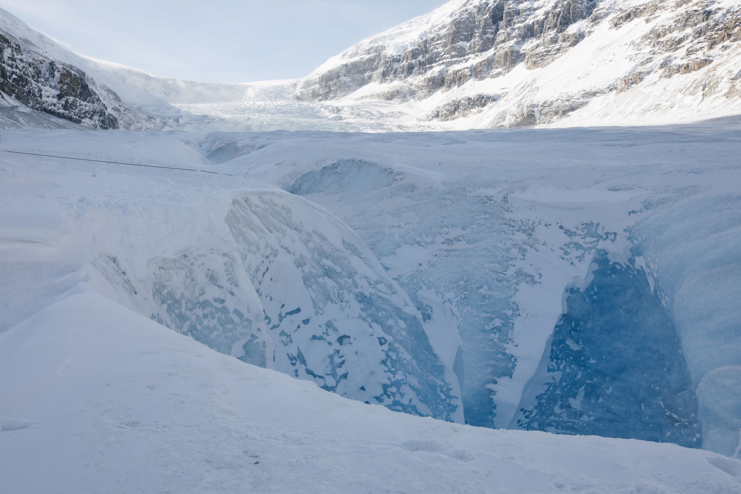 The blue ice of a moulin is in the foreground with snow on the sides and a glacier in the background.
