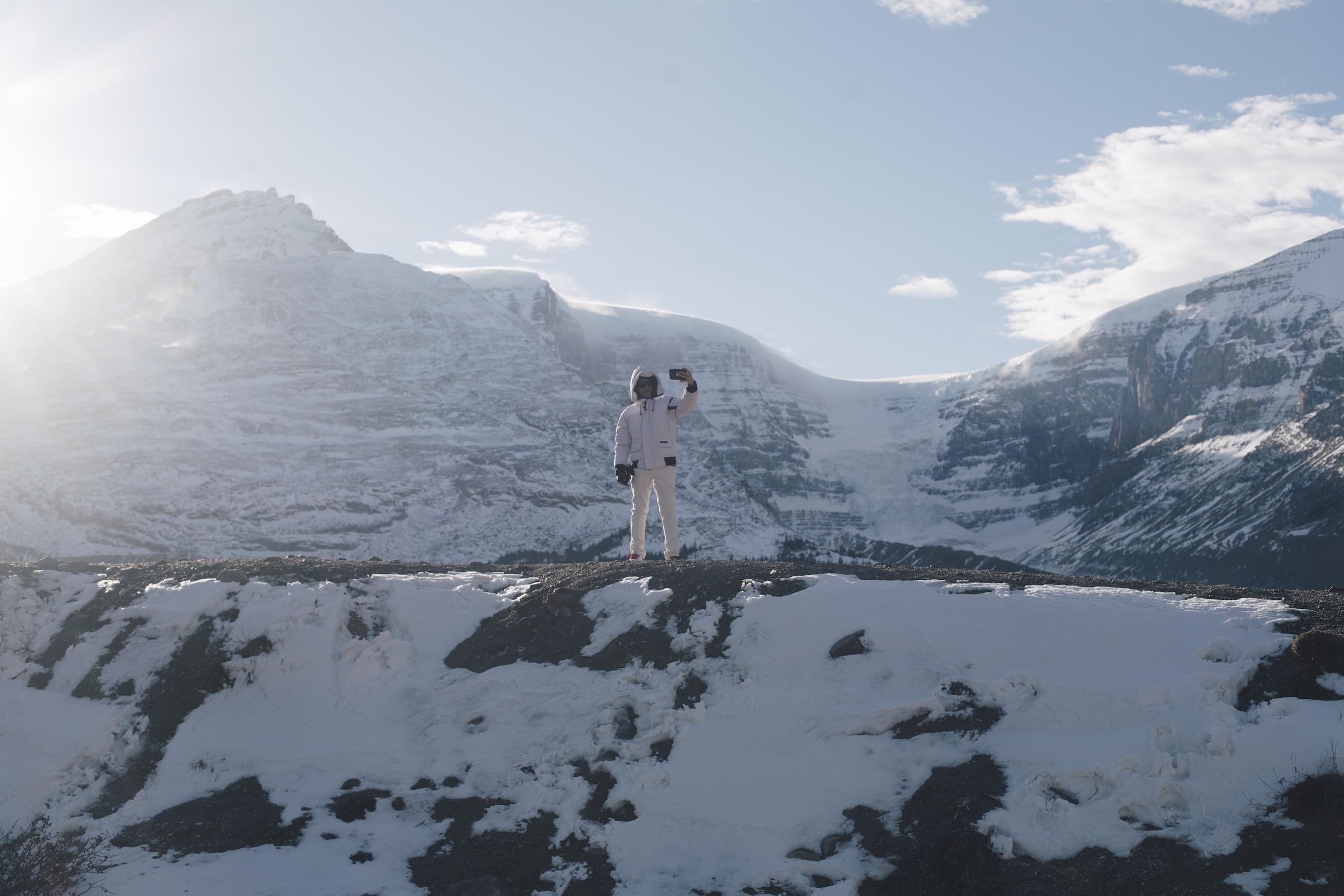 A tourist wearing all white holds up their phone to take a selfie in front of a glacier