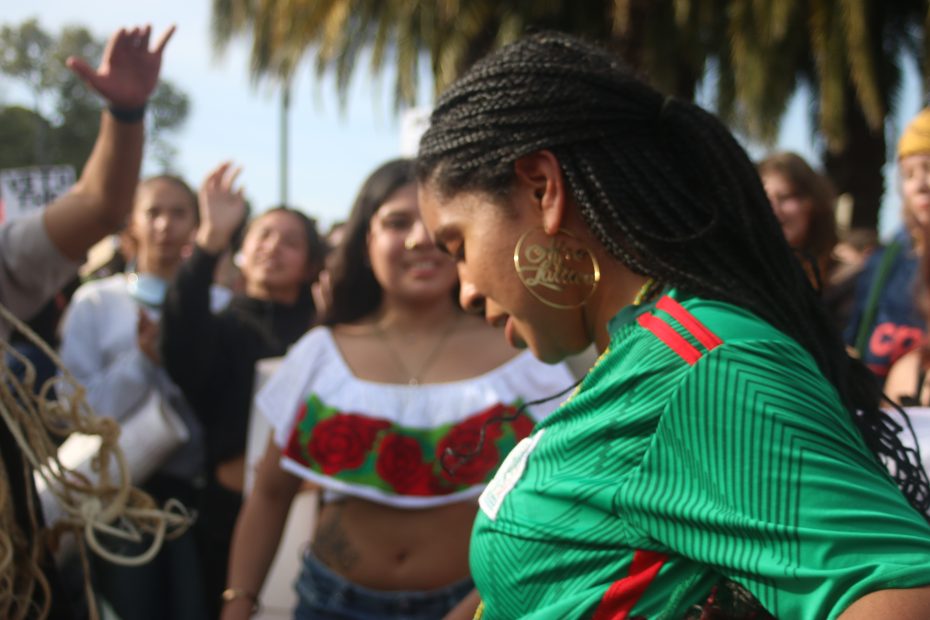 A woman with braided hair in a green jersey stands among a group of people outside during an ICE walkout; some are raising their hands, and trees are visible in the background.