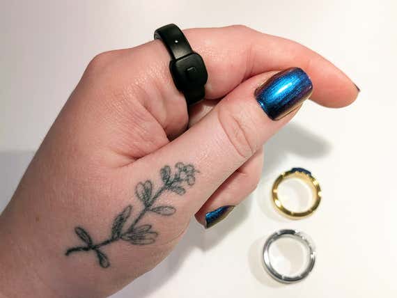 A close-up of a person's hand wearing the Pebble Index 01 smart ring on their index finger. The ring is a minimal, matte black band featuring a single physical button and a small microphone hole. On the table nearby, gold and silver versions of the ring are visible.