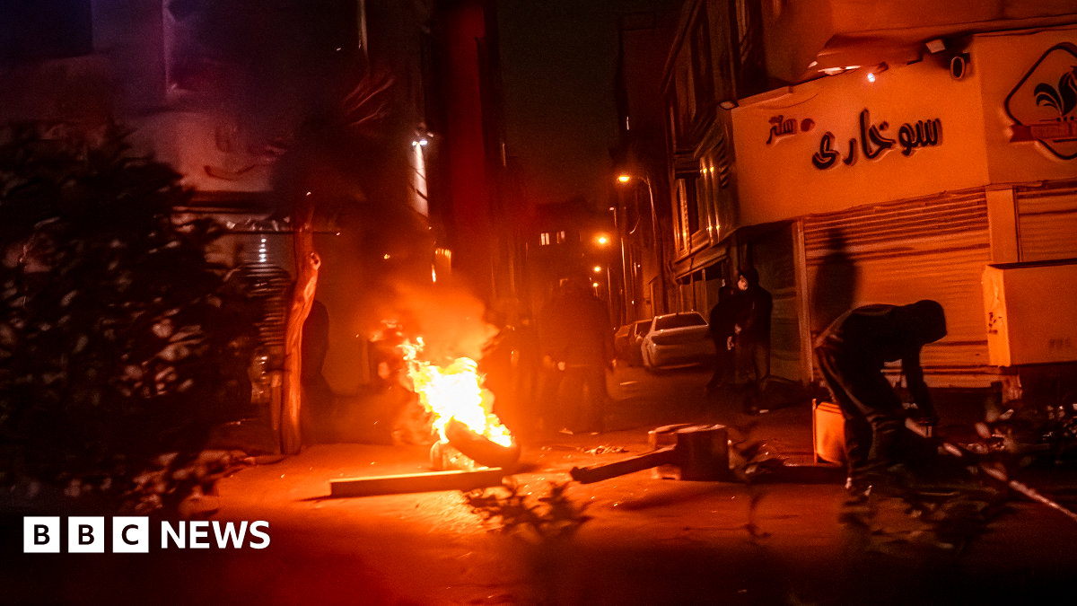 A scene of rioting in Iran - it is a wide shot of an urban street and a fire burns at a small barricade in the middle of the image, making the whole photo an orange-red colour. To the left of the camera is a bush, to the right a figure in black is hunched over a metal object. In the middle distance more black hooded figures can be seen.