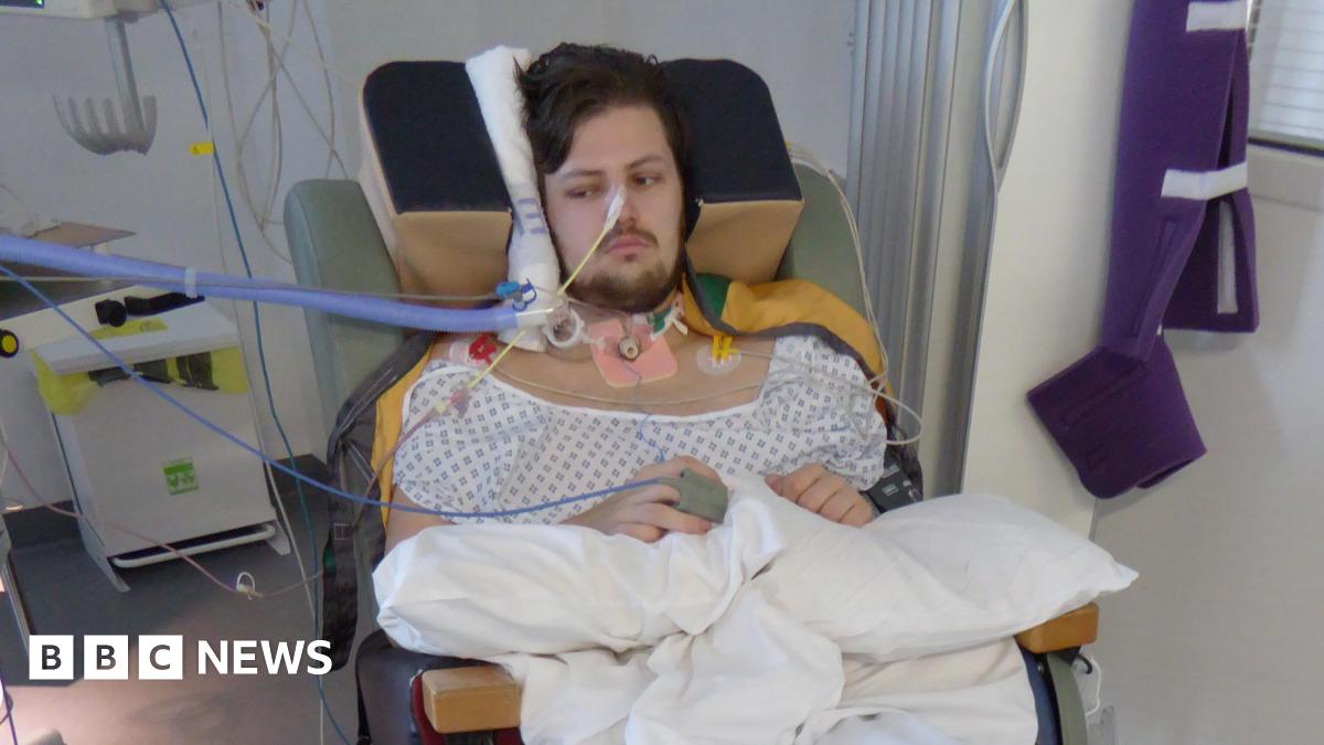 Nick Atherton sitting in a chair on a hospital ward. He has a tube coming out of his nose, one attached to his hand and his head and neck are supported by a variety of equipment. He is wearing a pale hospital gown and has a dark beard and short dark hair. His arms are resting on white pillows on his lap