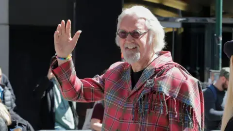 Getty Images Connolly wearing a red and green tartan plaid coat with fringed detailing on the shoulder, accessorized with a brooch and a colorful beaded bracelet, raising one hand in a wave. The background shows metal barricades and several people standing outdoors in a city setting.