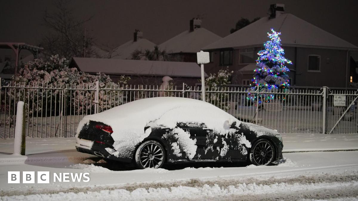 A black car covered in snow parked at the side of the road. The ground and houses behind the car are covered in snow. A tree decorated with Christmas lights can be seen in the garden behind the car.