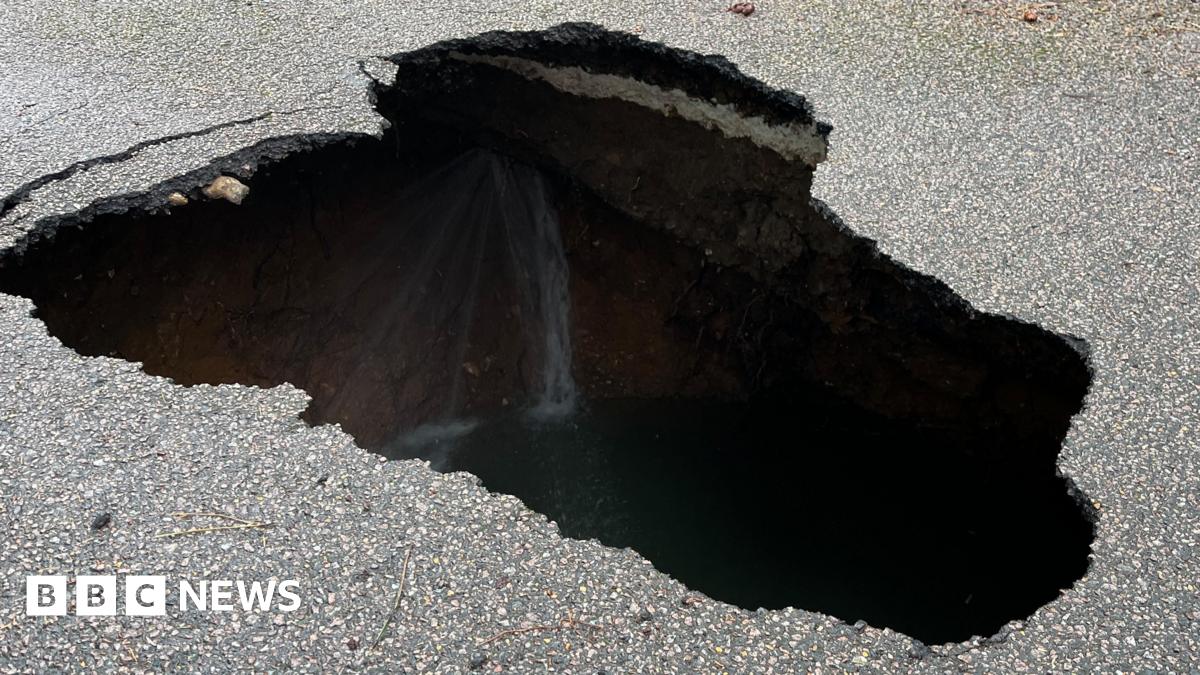 A residential street is cordoned off with blue-and-white police tape. A large hole or sinkhole is visible in the middle of the road.