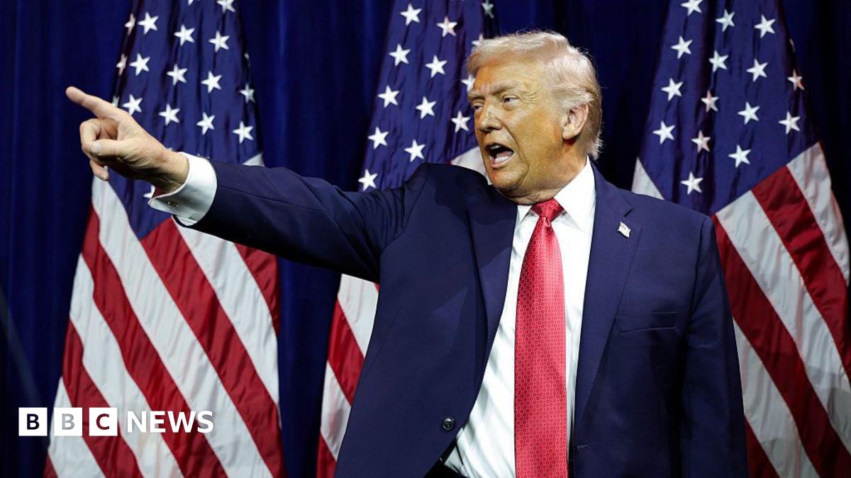 US President Donald Trump, wearing a blue suit and red tie, points with his finger in front of an American flag.