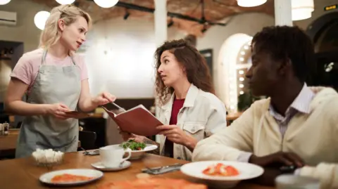 Getty Images/mediaphotos Disgruntled curly-haired young female sitting at restaurant table with boyfriend and reading menu while talking to waitress in modern loft cafe
